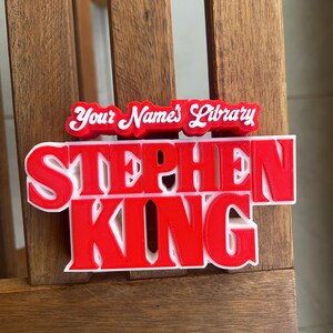 Może przedstawiać: Czerwona tabliczka 3D z nadrukiem z tekstem "Stephen King" i "Your Name's Library" w białych literach.