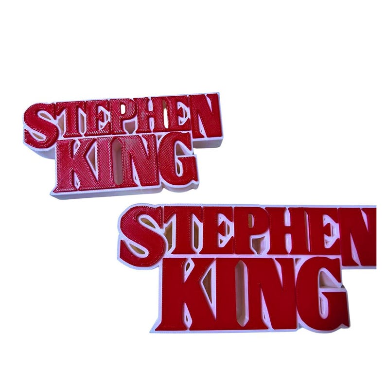 Może przedstawiać: Dwa czerwone znaki 3D wydrukowane z tekstem "Stephen King" białymi literami. Znaki znajdują się na białym tle.