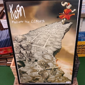 Op de afbeelding: Een ingelijste poster met een grunge rockband, Korn, en de woorden "Follow the Leader". De poster toont een cartoon afbeelding van kinderen die hinkelen op de rand van een klif. De klif brokkelt af en de kinderen dreigen te vallen. De poster is in een zwarte lijst en heeft een witte achtergrond.