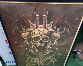 May include: Framed poster featuring a skull with a pentagram, surrounded by skulls, swords, and barbed wire. The poster has a dark color palette with red and green hues. The word "SLAYER" is printed at the bottom.