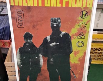May include: A framed poster with the text "TWENTY ONE PILOTS" in yellow letters. The poster features two figures in black clothing against a red and yellow background. Various symbols and the word "CLANCY" are also visible.