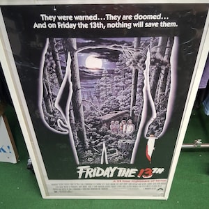 Può includere: Un poster cinematografico incorniciato per "Venerdì 13". Il poster presenta una scena di foresta con luna piena, una capanna e un gruppo di persone. Il titolo è in rosso e bianco, con lo slogan "Un incubo di terrore di 24 ore."