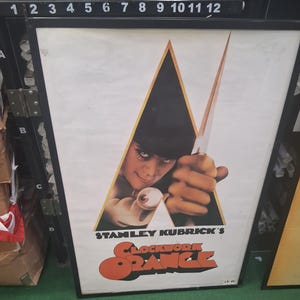 May include: A framed movie poster for Stanley Kubrick's A Clockwork Orange. The poster features a central triangular graphic containing a person's face and hand holding a knife, with the title text in bold orange letters at the bottom.