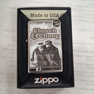 Peut inclure: Un briquet Zippo argent&eacute; avec une image en noir et blanc de Cheech & Chong. Le briquet est dans une bo&icirc;te noire et porte le logo Zippo en bas. Le texte "Made in USA" est visible.