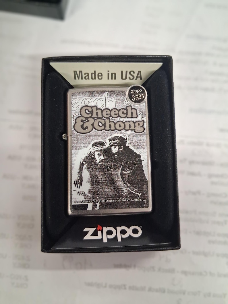 Peut inclure: Un briquet Zippo argent&eacute; avec une image en noir et blanc de Cheech & Chong. Le briquet est dans une bo&icirc;te noire et porte le logo Zippo en bas. Le texte "Made in USA" est visible.