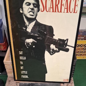 Può includere: Poster del film Scarface incorniciato con Al Pacino che tiene un fucile. Il poster presenta il titolo "SCARFACE" in lettere rosse e la frase "Say hello to my little friend..." in bianco. Il poster è incorniciato in nero.