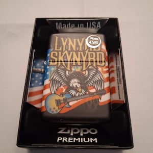 Puede incluir: Un encendedor Zippo Premium en una caja negra. El encendedor presenta un dise&ntilde;o con un &aacute;guila tocando la guitarra, las palabras "LYNY SKYNRYD" y un motivo de la bandera estadounidense. La caja est&aacute; etiquetada como "Made in USA".
