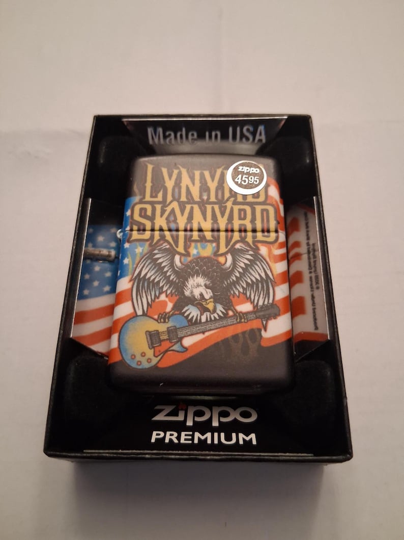 Puede incluir: Un encendedor Zippo Premium en una caja negra. El encendedor presenta un dise&ntilde;o con un &aacute;guila tocando la guitarra, las palabras "LYNY SKYNRYD" y un motivo de la bandera estadounidense. La caja est&aacute; etiquetada como "Made in USA".