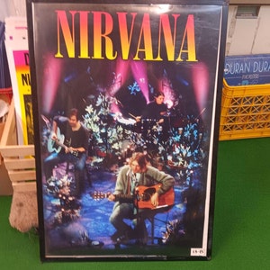 May include: A framed poster featuring the band Nirvana. The band members are performing on stage, with the band's name in large, colorful letters at the top. The stage is lit with pink and blue lights, and the band members are playing their instruments.