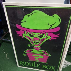 May include: Framed poster with a black background. It features a cartoon clown with green hair and a pink hat. The clown has a wide grin and a long tongue. The words "Riddle Box" are at the bottom.