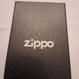 Puede incluir: Una caja rectangular negra con el logotipo blanco "Zippo" y un peque&ntilde;o icono de llama roja. La caja tiene una superficie texturizada y parece estar hecha de cart&oacute;n o un material similar. El logotipo de Zippo est&aacute; centrado en la parte superior de la caja.