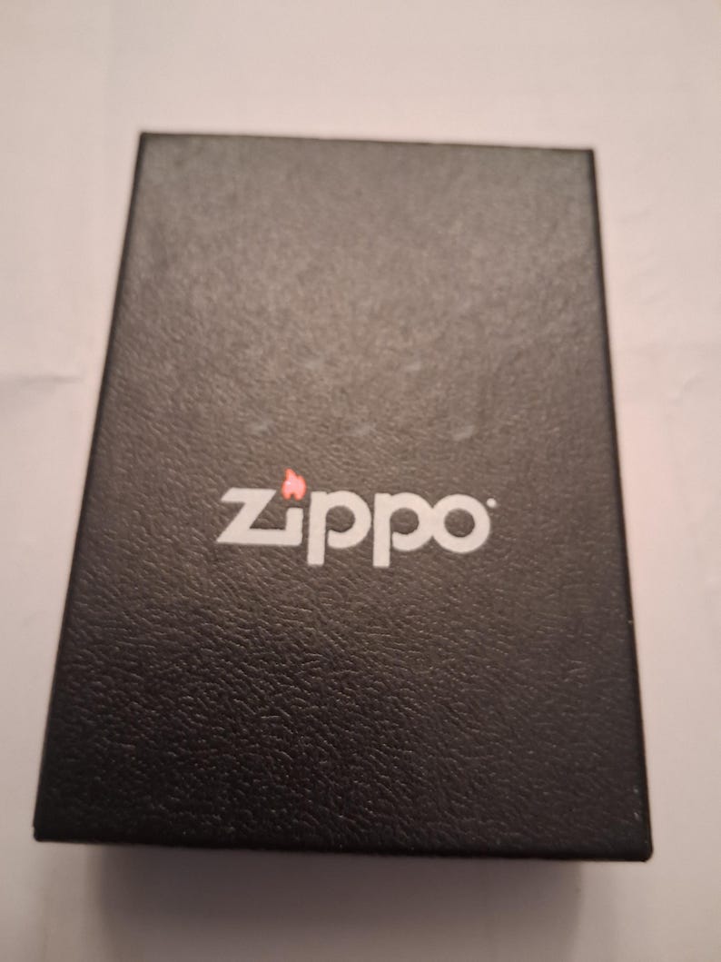 Puede incluir: Una caja rectangular negra con el logotipo blanco "Zippo" y un peque&ntilde;o icono de llama roja. La caja tiene una superficie texturizada y parece estar hecha de cart&oacute;n o un material similar. El logotipo de Zippo est&aacute; centrado en la parte superior de la caja.