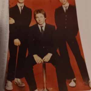 May include: Three men in black suits and white shirts pose for a photo. The man in the middle is sitting on a stool. The background is a solid red.