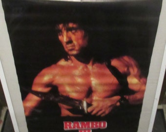 May include: A vintage movie poster for the film "Rambo III" featuring Sylvester Stallone as John Rambo. The poster has a black background with a close-up image of Rambo with his shirt off, holding a knife. The title "RAMBO III" is in red letters at the bottom of the poster.