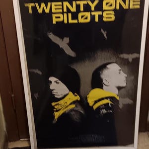 Puede incluir: Póster enmarcado de la banda Twenty One Pilots. El póster tiene un fondo negro con el nombre de la banda en letras amarillas. Dos miembros de la banda se representan de perfil, con detalles en amarillo. Aves blancas silueteadas en el fondo.
