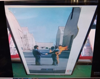 May include: A vintage poster featuring a black and white image of two men reaching out to each other with one man on fire. The background is a city street with buildings on either side. The poster has a blue and white rainbow design in the bottom right corner.