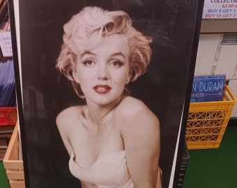 Könnte beinhalten: Ein schwarz-weiß gerahmtes Poster mit einem Porträt von Marilyn Monroe. Das Poster hat einen weißen Hintergrund und zeigt eine Nahaufnahme von Monroes Gesicht und Schultern. Sie trägt ein weißes trägerloses Kleid und hat ihre blonden Haare in einer weichen Welle gestylt. Das Poster ist unten links signiert.