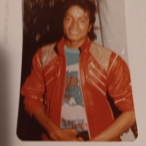 Puede incluir: Una foto de cerca de un hombre que lleva una chaqueta de cuero roja con un diseño de cremallera y una camiseta azul y blanca con la palabra "Impou" visible. El hombre tiene el pelo oscuro y sonríe.