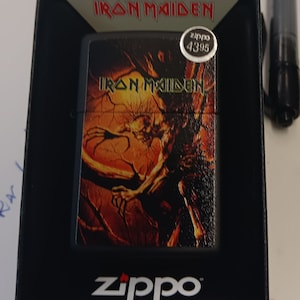 May include: A black Zippo lighter with a silver band around the top. The lighter has a graphic of a skeletal figure with flames on a black and orange background. The text "Iron Maiden" is printed in yellow on the lighter. The lighter is in a black box with the text "Iron Maiden" printed in white on the top.