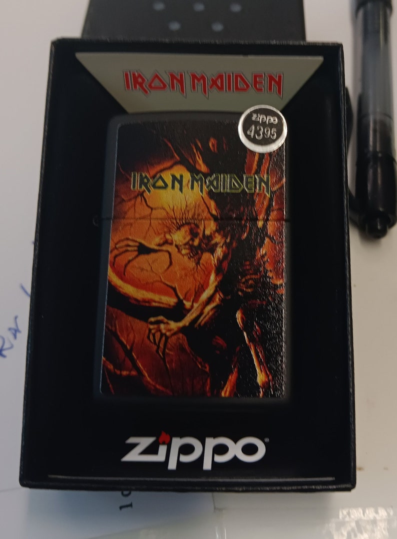 May include: A black Zippo lighter with a silver band around the top. The lighter has a graphic of a skeletal figure with flames on a black and orange background. The text "Iron Maiden" is printed in yellow on the lighter. The lighter is in a black box with the text "Iron Maiden" printed in white on the top.
