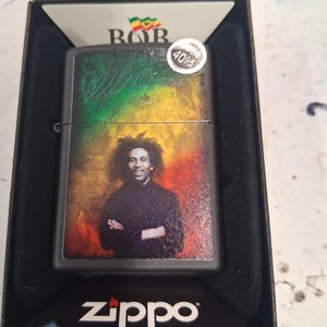 May include: A black Zippo lighter featuring a portrait of Bob Marley against a background of green, yellow, and red colors. The lighter is housed in a black display box with the Zippo logo printed at the bottom and a small sticker labeled 4095.