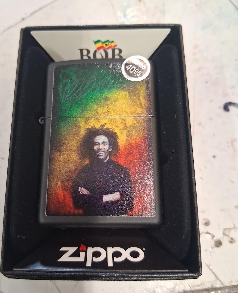 May include: A black Zippo lighter featuring a portrait of Bob Marley against a background of green, yellow, and red colors. The lighter is housed in a black display box with the Zippo logo printed at the bottom and a small sticker labeled 4095.
