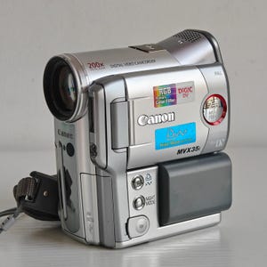 May include: A silver Canon MVX35i digital video camcorder featuring a 200x digital zoom lens, RGB primary colour filter, and DIGIC DV processor. The device includes a Mini DV slot, a Print Share button, and a Night Mode setting on the side panel.