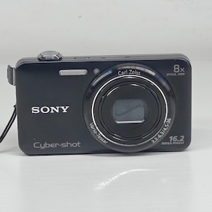 May include: A black Sony Cyber-shot digital camera featuring a Carl Zeiss Vario-Tessar lens with 8x optical zoom and 16.2 megapixels. The front of the device displays the Sony logo, the Cyber-shot branding, and technical lens specifications.