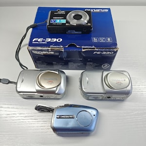 May include: A black Olympus FE-330 digital camera resting on its blue cardboard box, accompanied by two silver Olympus Camedia D-395 digital cameras and one light blue Olympus digital camera with an open battery compartment door.