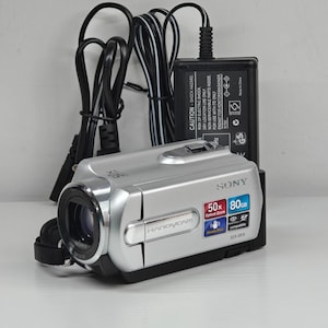 May include: A silver Sony Handycam DCR-SR15 camcorder with a 50x optical zoom lens and 80GB storage capacity. The device features a SteadyShot label and is accompanied by a black power adapter with a long cord and various printed safety warnings.