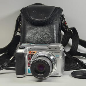 May include: A silver Kyocera Finecam M400R digital camera with a 10x optical zoom lens, featuring RTUNE branding and technical specifications printed on the front, accompanied by a black textured leather carrying case with an adjustable strap.
