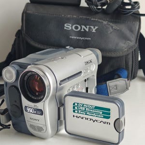 May include: A silver and dark grey Sony Digital8 Handycam video camera with a 20x optical zoom lens. The flip-out screen displays text reading DV IN/OUT, USB Streaming, and Easy Handycam. A black Sony carrying case is positioned behind the camera.