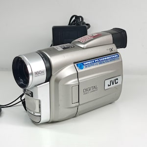 May include: A silver JVC digital video camera featuring a 160x digital zoom lens, a Mini DV port, and a Direct PC Connection label. The device includes a black power adapter with a cord and displays text indicating digital still output capabilities.