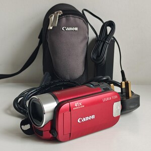 May include: A red Canon Legria FS306 camcorder with a 41x advanced zoom lens, accompanied by a black and grey Canon carrying case, a power adapter with a cord, and a three-pin electrical plug. The camcorder features a silver lens barrel.