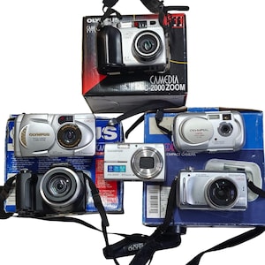 May include: A selection of Olympus digital cameras, some in their original packaging. The cameras are silver and black, with different zoom features. The boxes display the "CAMEDIA" and "C-2000 ZOOM" model names.