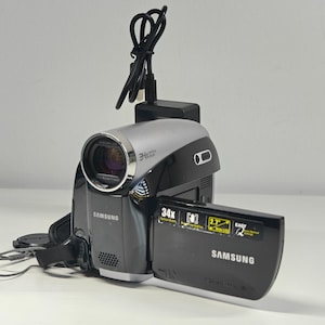 May include: A black and silver Samsung camcorder featuring a 34x optical zoom lens, a flip-out 6.86 cm wide LCD screen, and a black power cable connected to the top. The device displays the Samsung logo on the front and the side screen panel.