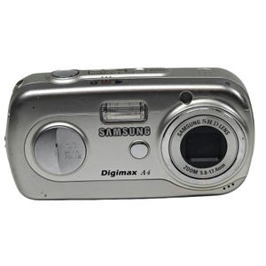 May include: A silver Samsung Digimax A4 digital camera featuring a 4.0 megapixels label, a flash unit, and a Samsung SHD lens with a 5.8-17.4mm zoom range. The top panel includes a power button and a shutter release button on the metallic body.