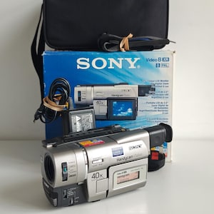 May include: A silver Sony Handycam Vision camcorder with a 40x digital zoom lens, a side-mounted LCD screen, and various control buttons. The camcorder is displayed in front of its original blue and white product box and a black carrying case.