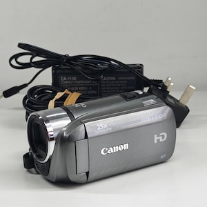 May include: A grey Canon Legria HF R205 HD camcorder with a 25x advanced zoom lens, featuring a silver metallic lens ring and a black power adapter with a cord. The device displays the Canon logo, HD branding, and technical specifications on its side.
