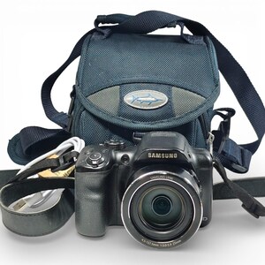 May include: A black Samsung digital camera with a lens marked 4.5-157.5mm 1:3.0-5.9 25mm, accompanied by a white USB cable and a dark blue camera bag featuring a silver oval logo with a fish graphic and a shoulder strap.