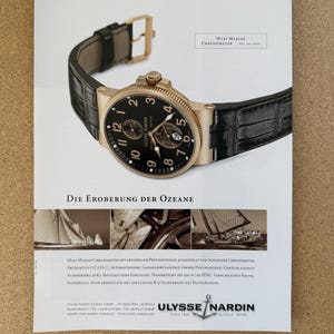 May include: A gold-toned Ulysse Nardin Maxi Marine Chronometer watch with a black face and leather strap. The watch has a date display and is set against a white background with German text. A luxury watch.