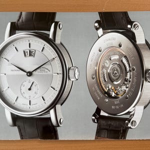 Puede incluir: Un reloj de pulsera plateado con una correa de cuero negro, que muestra la parte delantera y trasera. La parte delantera presenta una ventana de fecha y un pequeño dial de segundos. La parte trasera revela el mecanismo. El reloj está marcado "STAINLESS STEEL" y "MADE IN GERMANY".