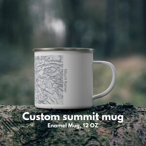 Könnte beinhalten: Weißer Emaille-Becher mit silbernem Rand, mit einem topografischen Kartendesign und dem Text "Mount Rainier". Der Becher hat auch den Text "Custom summit mug" und "Emaille-Becher, 340 ml."