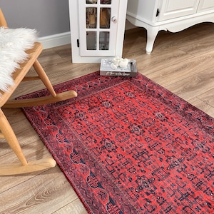 May include: A rectangular area rug featuring a vibrant red base color with intricate dark blue and black geometric patterns and floral motifs. The rug has a detailed border design and a flat-woven texture, covering a portion of the wooden floor.