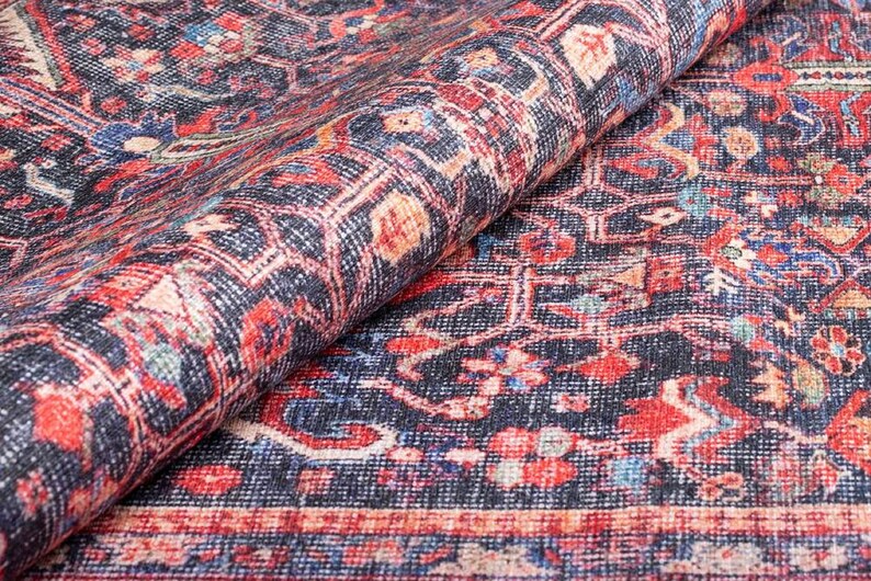 May include: A folded area rug featuring a dark navy blue background with an intricate pattern of red, cream, and light blue floral and geometric motifs. The textile displays a dense, woven texture with a visible border along the bottom edge.