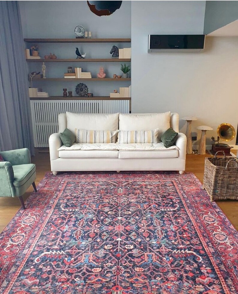 May include: A large rectangular area rug featuring a dark blue base with intricate floral and geometric patterns in shades of red, pink, and light blue. The rug is positioned in a room with a white sofa, a green armchair, and a wicker basket.