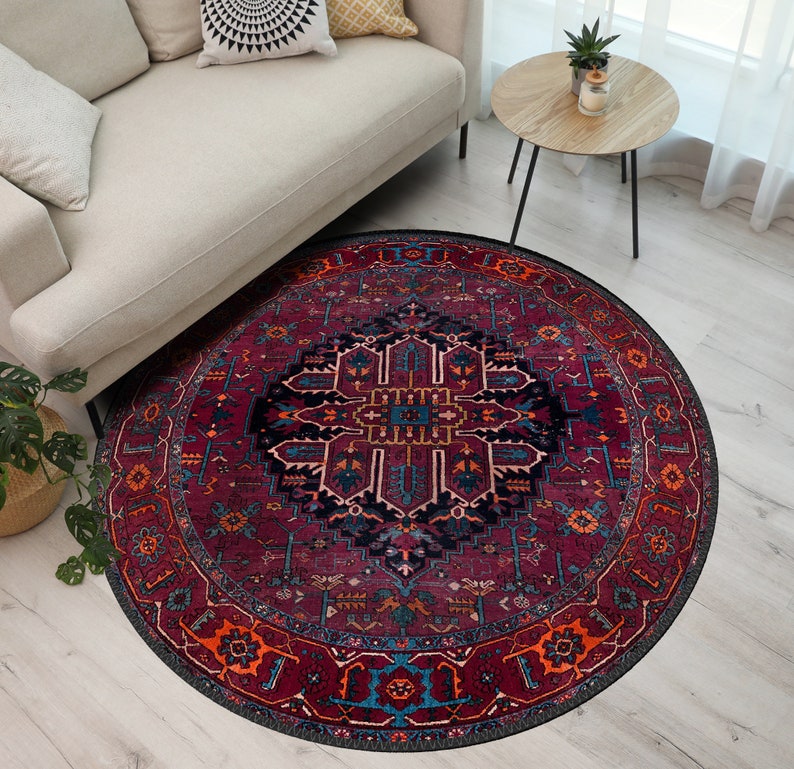 May include: A round area rug with a traditional Persian design. The rug features a deep burgundy background with intricate patterns in navy, orange, and teal. The rug is placed on a light wood floor, partially under a beige sofa.