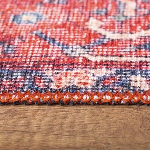 May include: A woven rug featuring a detailed pattern in shades of red, navy blue, and cream. The edge of the rug is finished with a thick, orange overlock stitch, creating a distinct border along the side of the textile material.