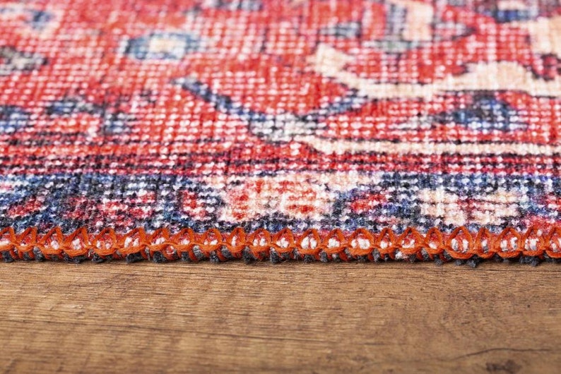 May include: A woven rug featuring a detailed pattern in shades of red, navy blue, and cream. The edge of the rug is finished with a thick, orange overlock stitch, creating a distinct border along the side of the textile material.