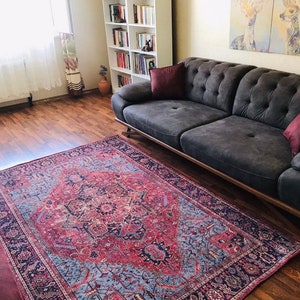 May include: A red, blue, and black patterned rug with a detailed design. The rug is laid out on a hardwood floor in a living room setting with a dark gray sofa and a white bookshelf in the background.
